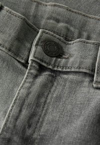 Gråt denimstof med synlig tekstur, med en sort knap præget med "TWO SOONS JEANS" og kontrasterende syninger. Nærbillede.