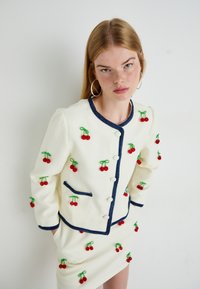 Mujer joven con falda blanca y chaqueta abotonada con ribete azul marino y bordado de cerezas rojas, de pie con las manos en los bolsillos frente a una pared lisa.