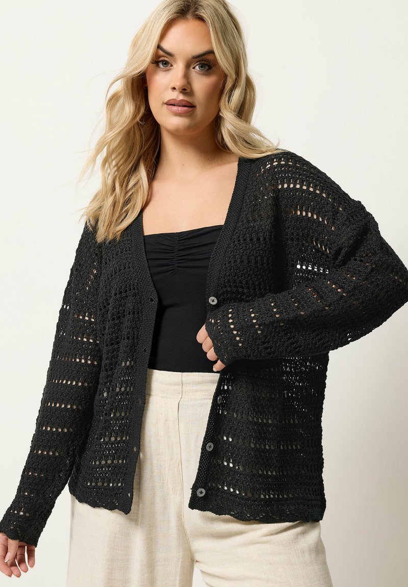 Femme blonde portant un cardigan noir en crochet par-dessus un haut noir et un pantalon crème taille haute, debout devant un fond uni.