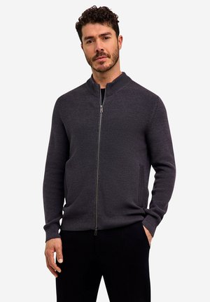 Mann mit kurzen lockigen Haaren, der einen dunkelgrauen Reißverschluss-Pullover und schwarze Hosen trägt, steht mit einer Hand in der Tasche vor neutralem Hintergrund.