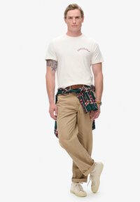 Camiseta de algodón beige con el logo rojo de "Superdry", pantalones de pana color camel y camisa de cuadros verde anudada a la cintura; zapatillas blancas.