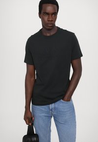 Černé bavlněné tričko s vyvýšeným logem "A|X" a "ARMANI EXCHANGE." Model drží černou tašku. Kombinováno se světle modrými džínami.