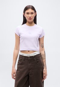 Ανοιχτό μωβ κοντομάνικο τοπ, με ανάγλυφο λογότυπο "CK". Φορεμένο με καφέ παντελόνι ψηλόμεσο και λευκή λεπτομέρεια στη μέση.