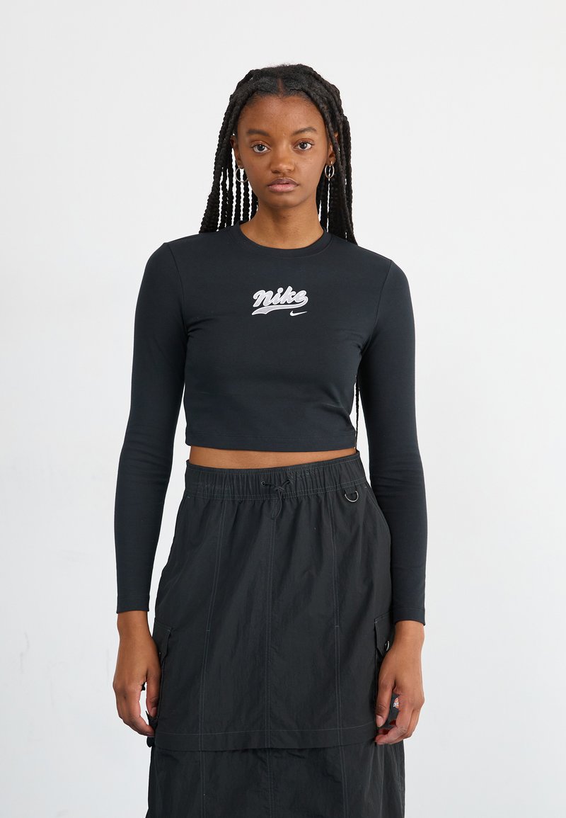 Fekete, crop hosszúujjú póló fehér "Nike" logóval. Fekete, zsinóros derekú szoknyával viselve, amely zsebekkel és réteges kialakítással rendelkezik.