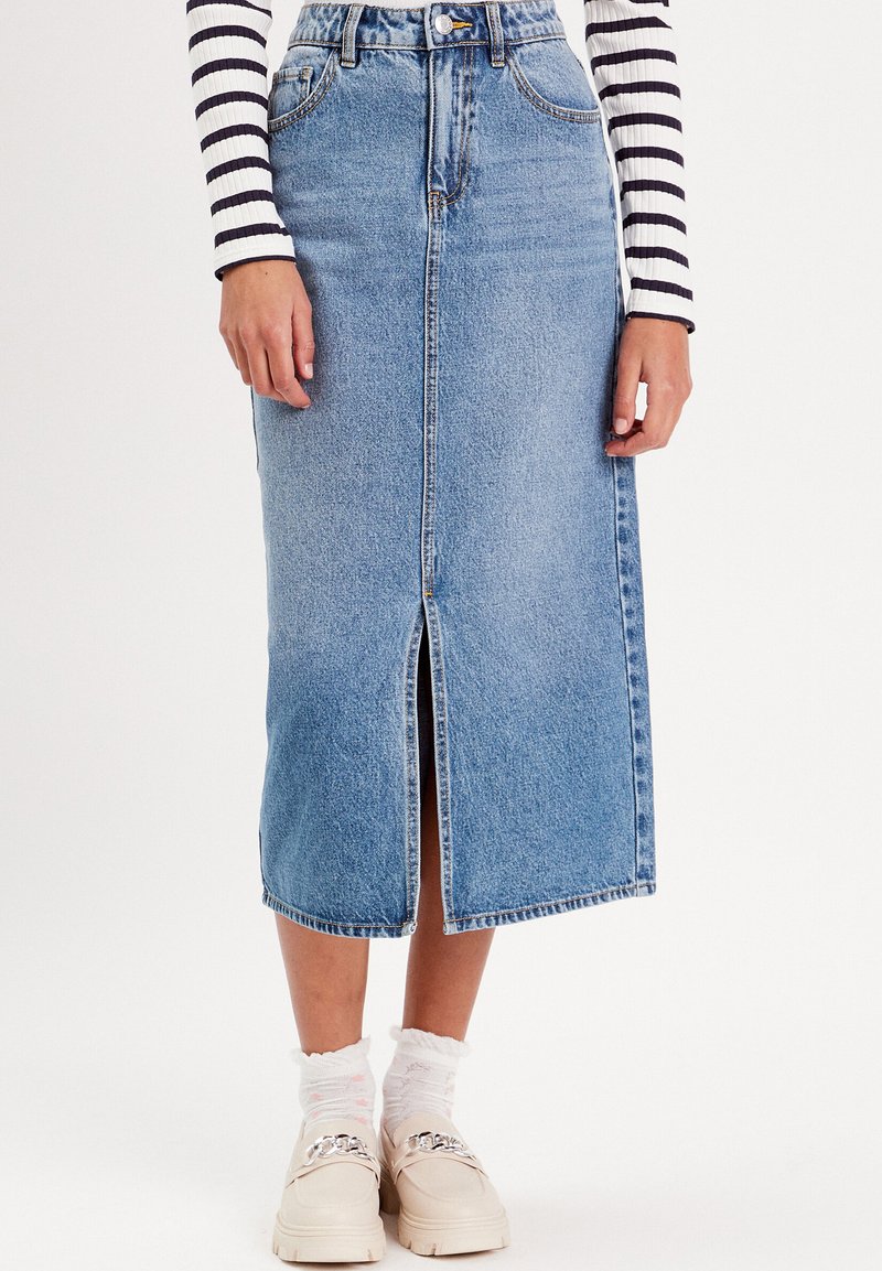 Jupe en denim bleu clair, longueur genou avec une fente à l'avant. Équipée de passants de ceinture et d'un design à cinq poches, assortie de chaussettes blanches et de chaussures claires.