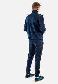 Marineblauer Trainingsanzug mit einer Jacke mit hohem Kragen, elastischen Bündchen und passenden Hosen mit schwarzen Seitenstreifen. Kombiniert mit marineblauen Sportschuhen.