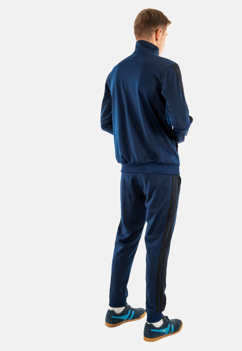 Survêtement bleu marine avec une veste à col haut, des poignets élastiques, et un pantalon assorti avec des rayures noires sur les côtés. Association avec des chaussures de sport bleu marine.