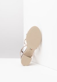 Sandal con tacco alto nuda con cinturini metallici, suola a texture liscia e etichetta del marchio embossata; esposizione inclinata mostra il design della parte inferiore.