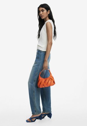 Orange woven handbag with a curved shape, small handle, and visible stitching; model wears a white top, blue jeans, and blue high-heeled sandals.