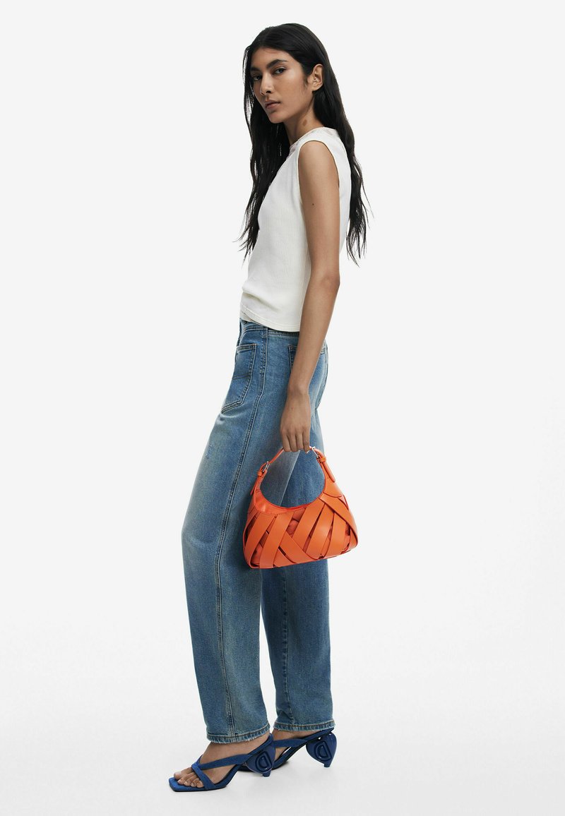 Orange woven handbag with a curved shape, small handle, and visible stitching; model wears a white top, blue jeans, and blue high-heeled sandals.