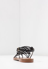 Sandale noire à brides avec un design en cuir tressé, semelle en bois plate et petit détail de boucle dorée à la cheville.