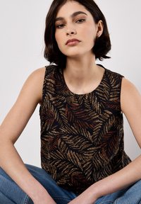 Ærmeløs top med en rund udskæring. Sort stof med et mønster af gyldne og rustfarvede bregner. Modellen sidder iført blå jeans foran en neutral baggrund.