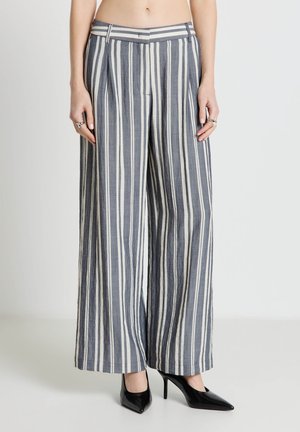 Frau trägt hoch taillierte, breit geschnittene Hose mit vertikalen blau-weißen Streifen, kombiniert mit schwarzen spitzen High Heels.