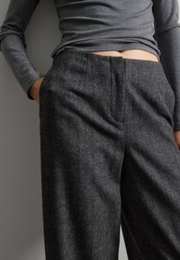 Haut à manches longues gris associé à un pantalon en chevrons gris foncé présentant des plis à l'avant et une coupe décontractée, les mains dans les poches.