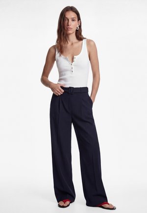 Kobieta ubrana w biały, bezrękawnik zapinany na guziki, granatowe, szerokie spodnie z wysokim stanem i paskiem oraz czerwone klapki, stoi z jedną ręką w kieszeni.