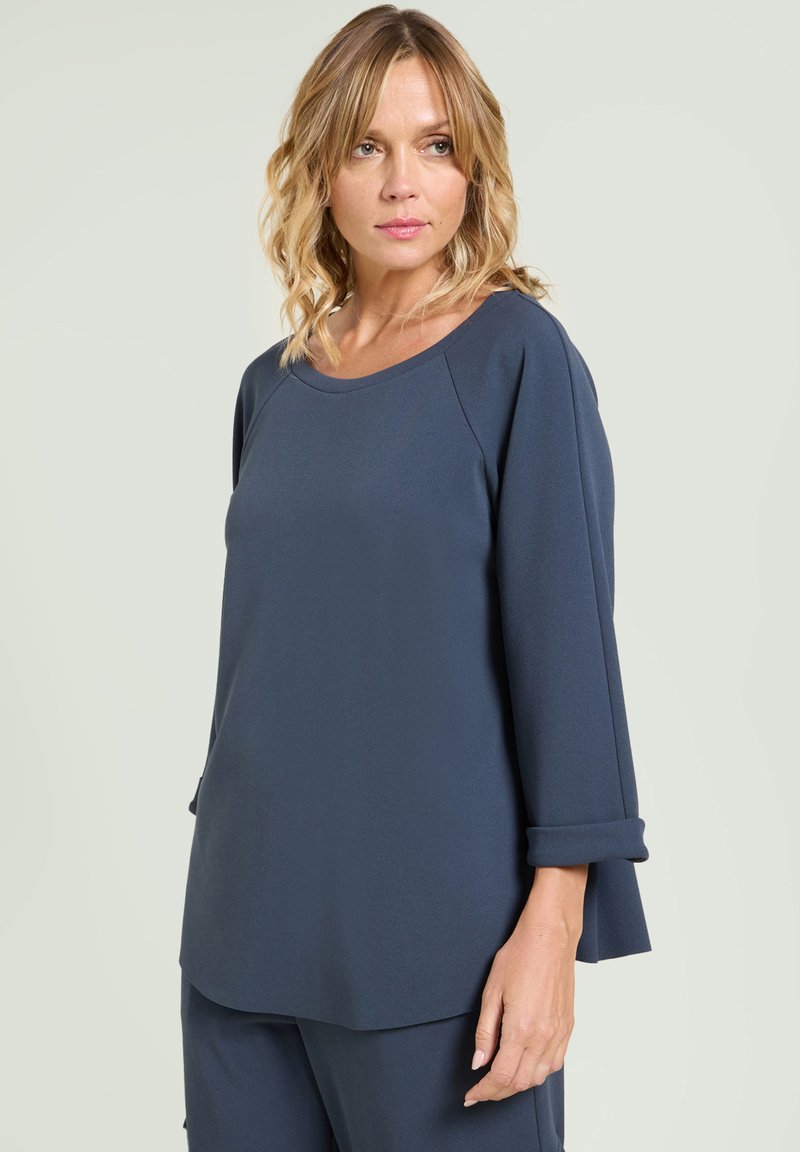 Mujer con cabello rubio hasta los hombros, vistiendo una blusa larga de manga larga suelta y oscura de color azul, y pantalones a juego, mirando ligeramente de lado.