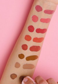 Muestras de 12 colores de labios aplicadas en un brazo, mostrando una gama desde rojos hasta marrones sobre un fondo rosa. Algunos tienen acabados brillantes.
