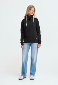 Felpa nera con un alto collo arricciato, lacci e dettagli marroni. Abbinata a jeans azzurri chiari e sneakers grigie.