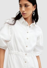 Weißes Denim-Kleid mit kurzen Puffärmeln, einer Knopfleiste und einem elastischen Bund. Verfügt über goldfarbene Knöpfe und einen kragenähnlichen Halsauschnitt.