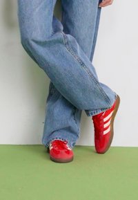 Rde teniske z belimi črtami, iz semiša in mrežne tkanine, zaobljenega prsta, gumijastim podplatom, oblečene z svetlo modrimi zvonastimi kavbojkami na zeleni tleh.