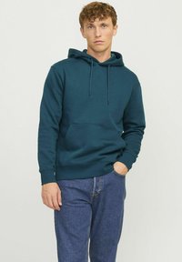Türkisfarbener Kapuzenpullover mit einer Vordertasche, Zugbandkapuze, gerippten Bündchen und Saum. Getragen mit blauen Denim-Jeans im lässigen Schnitt.