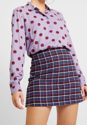 Femme portant une chemise lavande à boutons avec de grands pois rouges, associée à une jupe à carreaux bleue et rouge taille haute.