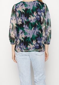 Blusa trasparente con un motivo floreale multicolore in verde, viola e nero. Maniche lunghe a sbuffo con polsini elastici.