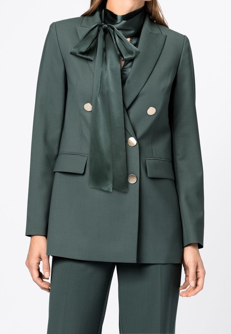 Femme portant un blazer croisé vert foncé à boutons dorés et un pantalon assorti, associé à un chemisier en satin vert foncé avec un nœud papillon.