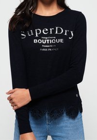 Marinblå långärmad topp med "SuperDry"-logotyp. Har spetsdetaljer vid fållen. Mjuk tygkvalitet med avslappnad passform. Bärs med ljusblå jeans.