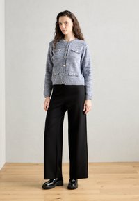 Cardigan bleu tricoté avec des boutons dorés et deux poches avant, associé à un pantalon noir ample et des chaussures noires épaisses.