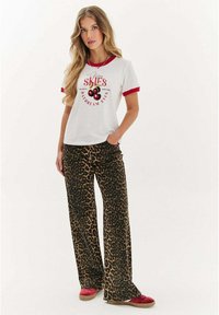 Bílý tričko s červeným límcem a grafickým textem. Hnědé kalhoty s širokým leopardím vzorem. Červené slip-on boty. Měkká tkanina s uvolněným designem.