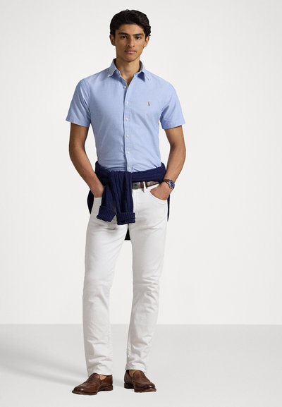 Chemise bleu clair à manches courtes avec boutons, pantalon blanc, pull marine noué à la taille, mocassins marron. Design simple avec peu de détails.