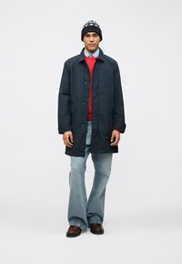 Námornícky ľahký vonkajší kabát, červený sveter, modrá košeľa, svetlé rifľové zvonové nohavice, hnedé topánky, pletená čierna a sivá čiapka so vzorom.