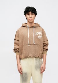 Rudas, tekstūruotas džemperis su kapišonu, priekyje kišenė, balti raišteliai ir logotipo detalės. Derinamas su šviesių spalvų kelnėmis, turinčiomis tamsias akcentus.