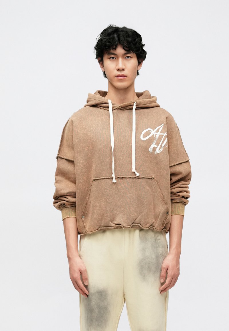 Rudas, tekstūruotas džemperis su kapišonu, priekyje kišenė, balti raišteliai ir logotipo detalės. Derinamas su šviesių spalvų kelnėmis, turinčiomis tamsias akcentus.