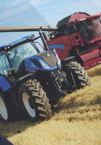 Blauer Traktor mit großen, robusten Reifen, vorne montierter Ausstattung und sichtbarem Fahrer. Roter Mähdrescher im Hintergrund. Stoppelfeld.
