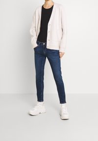 Cardigan de punto beige claro con botones, camiseta negra debajo, jeans oscuros ajustados y zapatillas deportivas blancas con suela gruesa. Sin patrones ni acentos.