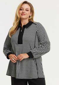 Damenoberteil mit Hahnentrittmuster in Schwarz und Weiß, mit einem Viertelreißverschluss und schwarzem Kragen. Lange Ärmel mit schwarzen Akzenten an den Bündchen.