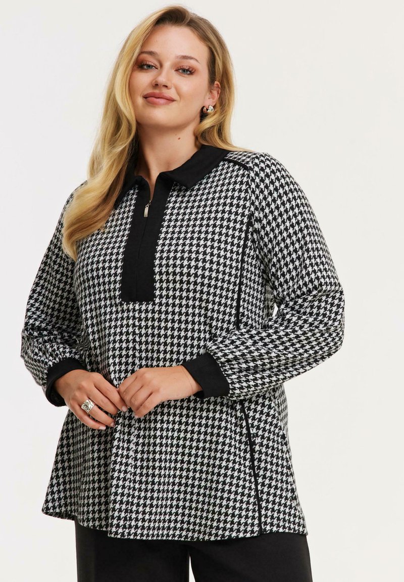 Damenoberteil mit Hahnentrittmuster in Schwarz und Weiß, mit einem Viertelreißverschluss und schwarzem Kragen. Lange Ärmel mit schwarzen Akzenten an den Bündchen.