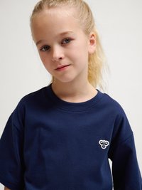 Lyshåret pige med hestehale iført en mørkeblå T-shirt med et lille hvidt og blåt biemblem på venstre bryst, kigger ind i kameraet.