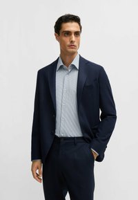 Veste marine sur une chemise rayée bleu clair. La veste a deux boutons et un col cranté. La tenue est complétée par un pantalon assorti.