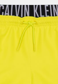 Neon žute kupaće gaće s elastičnim crnim pojasom na kojem stoji "CALVIN KLEIN" u srebrnoj boji. Uključuju i šnalu za podešavanje.