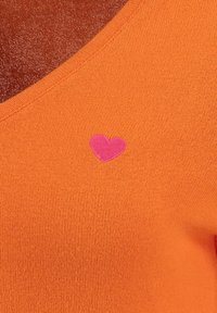 Orange gestricktes Oberteil mit V-Ausschnitt und einem kleinen rosa Herzsymbol auf der Brust. Strukturierter Stoff mit glatter Oberfläche.