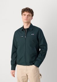 Donkergroene ritsjack met een kraag, zijzakken en een klein logo op de borst. Heeft een gladde textuur en een getailleerd ontwerp.