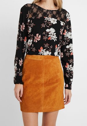 Sort blomstret blondefelt overdel kombineret med en sennepsfarvet ruskind mini nederdel, vist på en stående model mod en ensfarvet baggrund.