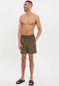 Costume da bagno verde oliva con vita elasticizzata, cordino nero e tasca laterale; indossato con sandali neri.