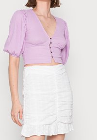 Top corto lavanda con maniche a sbuffo e scollatura profonda a V, caratterizzato da bottoni decorativi. Abbinato a una mini gonna bianca texturizzata con volant.