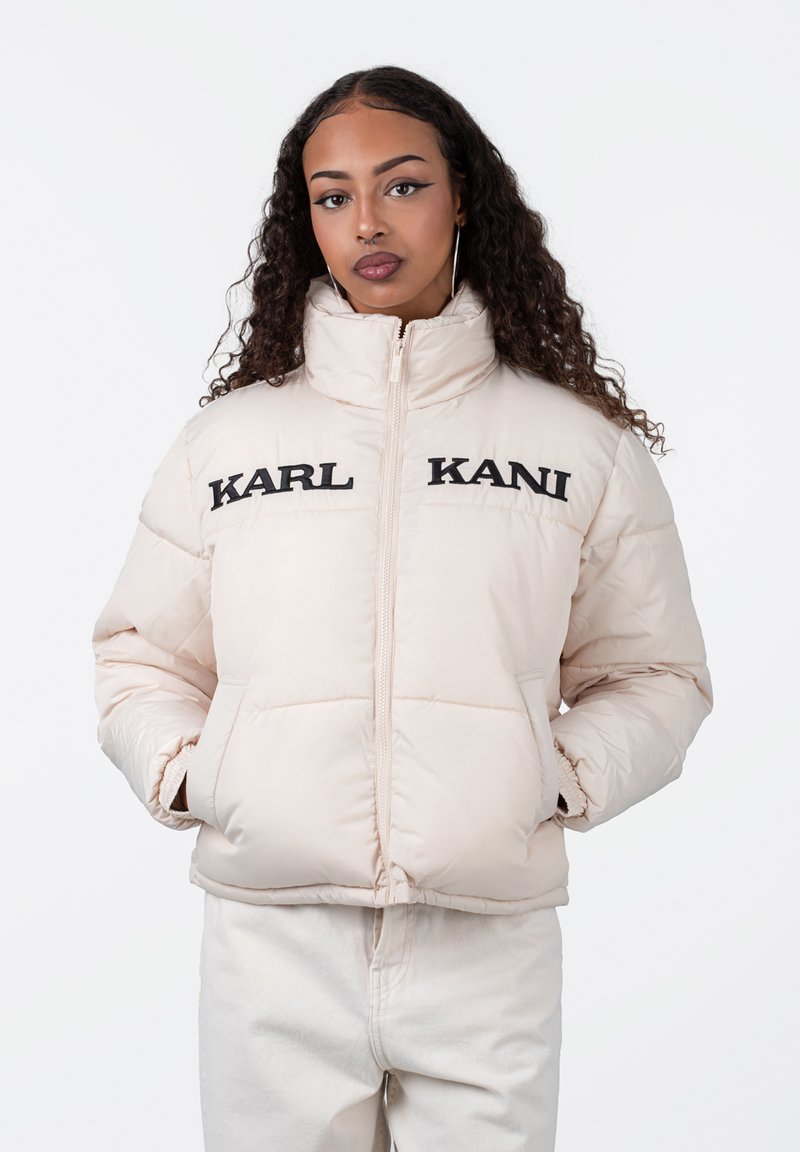 Lys beige puffer-jakke med høy krage, med «KARL KANI» brodert i svart på brystet. Glatt tekstur, glidelås og sidelommer.