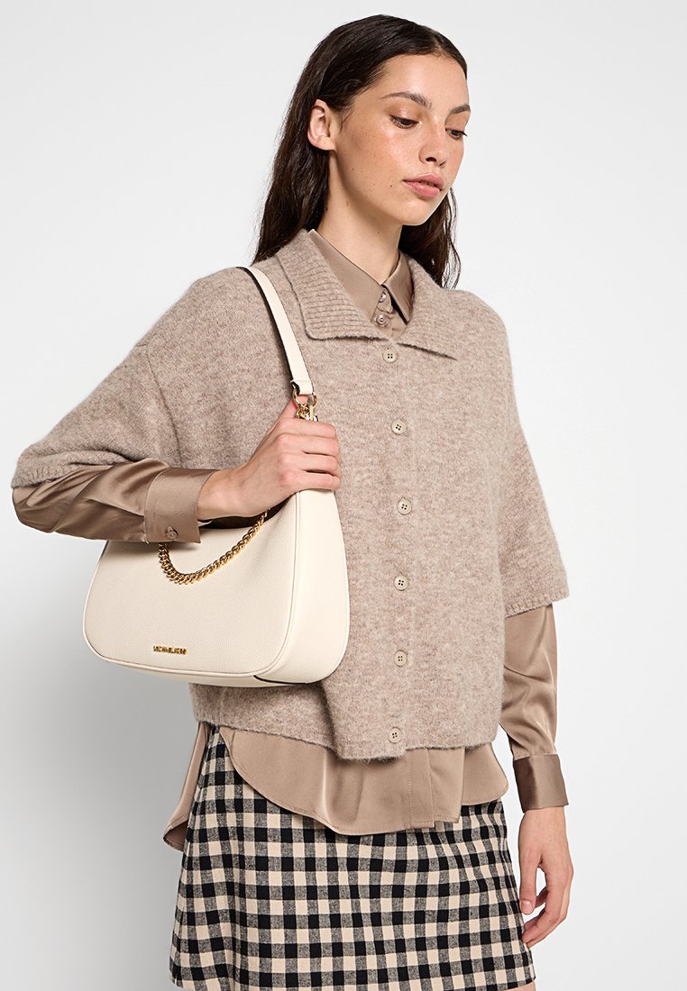 Sweat à col et boutons de couleur beige clair, coupé au niveau du ventre, associé à une chemise en satin marron clair et une jupe à carreaux noir et blanc. Sac à main blanc avec un accent en chaîne.