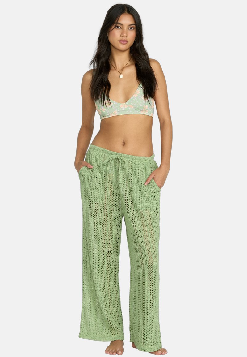 Pantaloni in rete verde chiaro con vita regolabile e tasche laterali, abbinati a un top reggiseno con fantasia floreale. Stilizzati per comfort e traspirabilità.